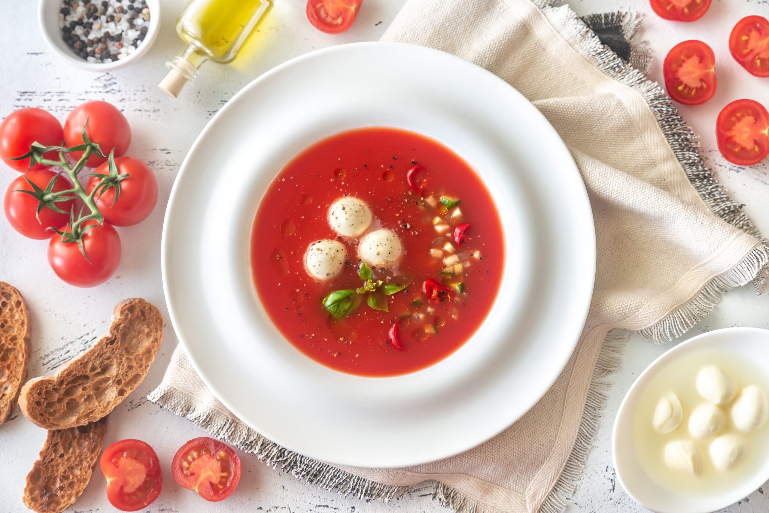 Porzione di gazpacho con ingredienti freschi disposti intorno e fette di pane.