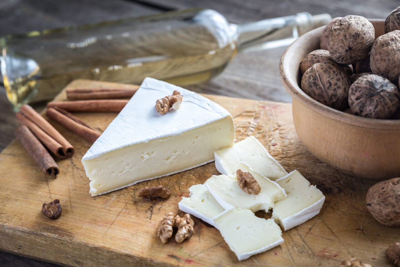Fette di formaggio brie, gherigli di noci e stecche di cannella appoggiati su un vassoio di legno insieme a un contenitore con noci in guscio, sullo sfondo c'è una bottiglia di vino bianco posta in orizzontale